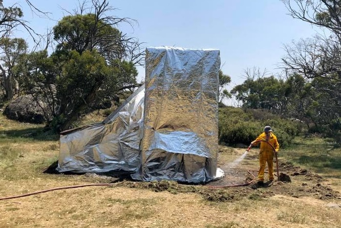A firefighter uses a hose to douse the land around the base of a hut entirely covered in foil.