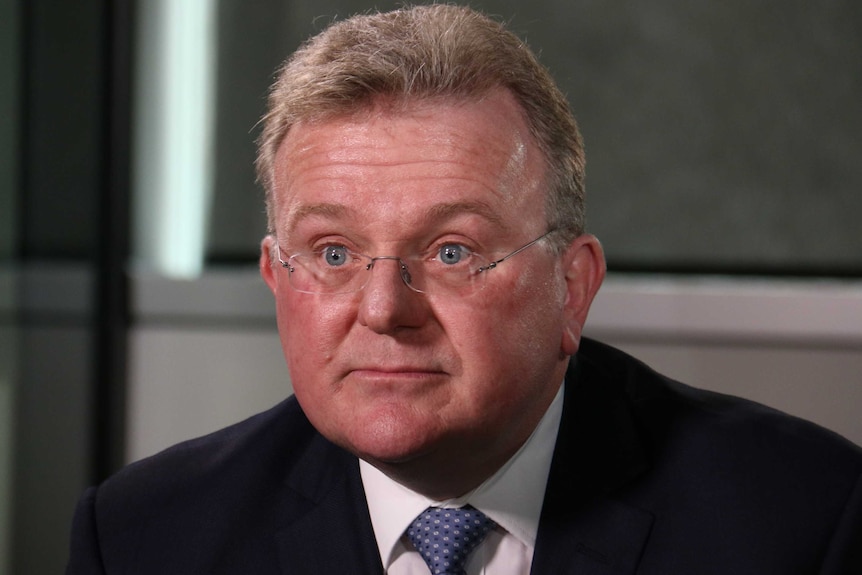 A bespectacled man greying at the temples, formally dressed for a television interview.