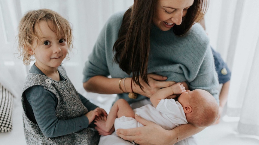 The mother breastfeeds her baby while the toddler watches a story about the cost of childbirth.