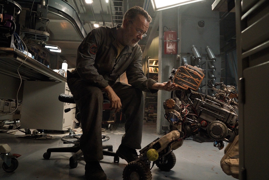 Middle-aged man with grey beard and glasses sits at a work desk in an industrial workshop space and tinkers with a robot.