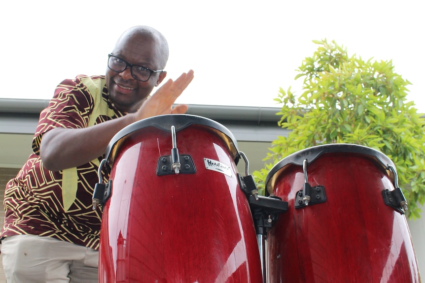 Allan Chidziva wears a yellow and brown African shirt and plays two red bongo drums.