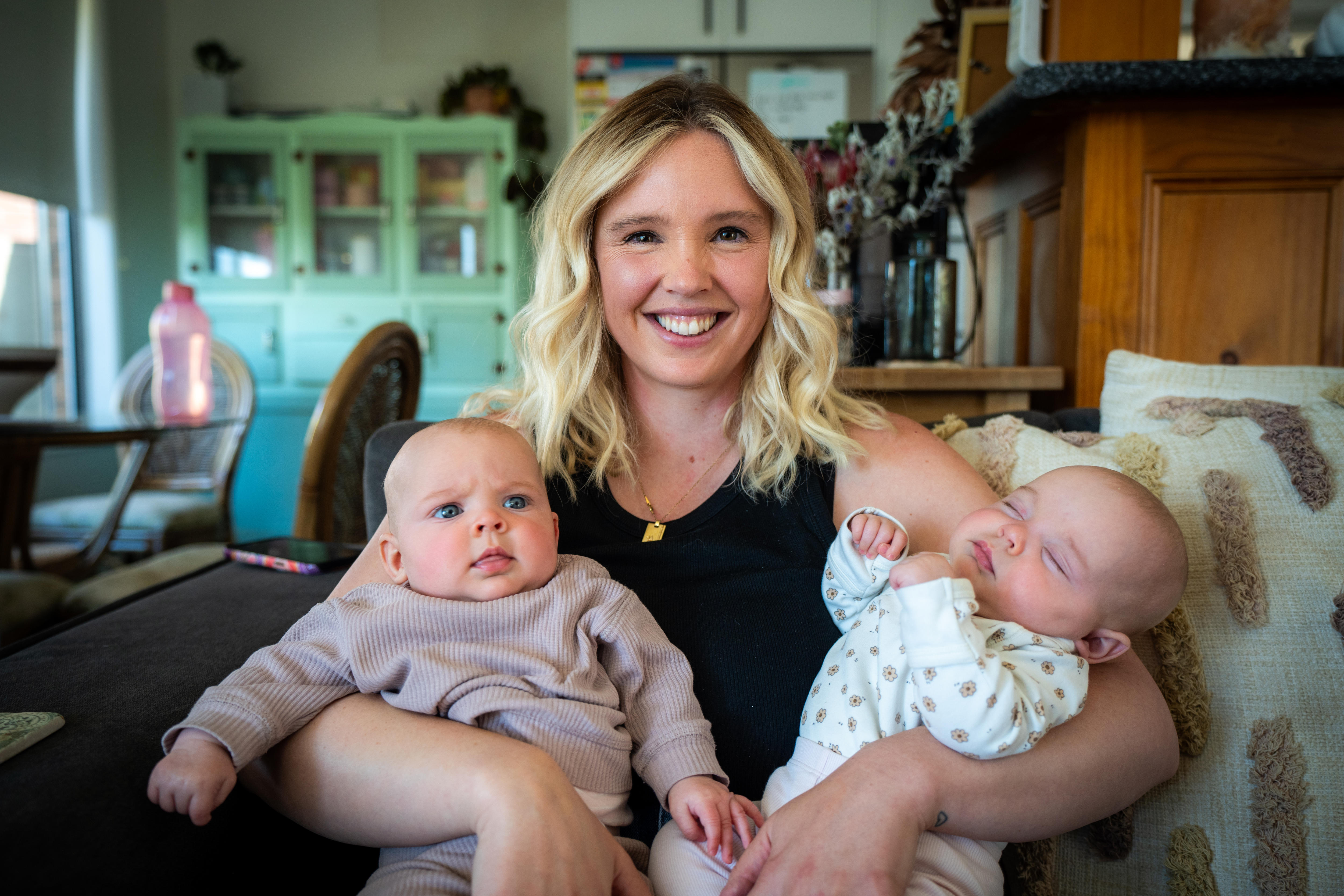 A smiling woman sits on the couch holding two babies, one in each arm