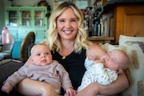 A smiling woman sits on the couch holding two babies, one in each arm