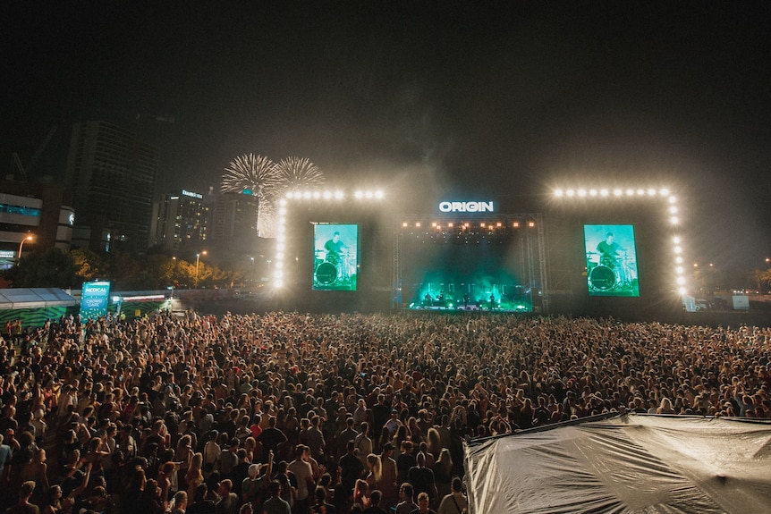 A wide shot of a crowd in front of a stage with fireworks in the background.