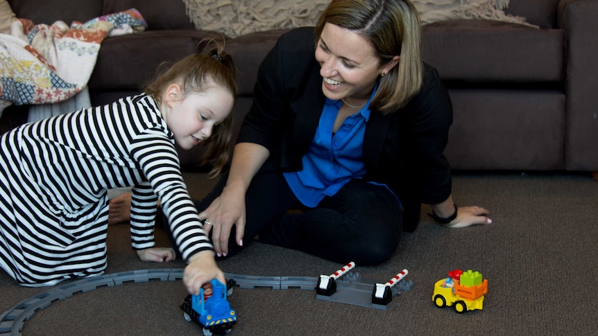A woman and a young girl play with a toy train on the floor in front of a couch.
