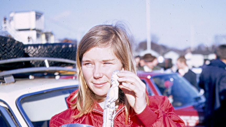 A young blonde women stands next to a rally car with crowds in the background.