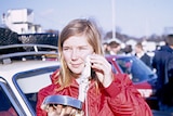 A young blonde women stands next to a rally car with crowds in the background.