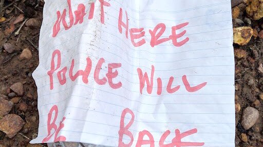 A message written on a piece of paper and taped to a plastic water bottle reads 'WAIT HERE POLICE WILL BE BACK'.