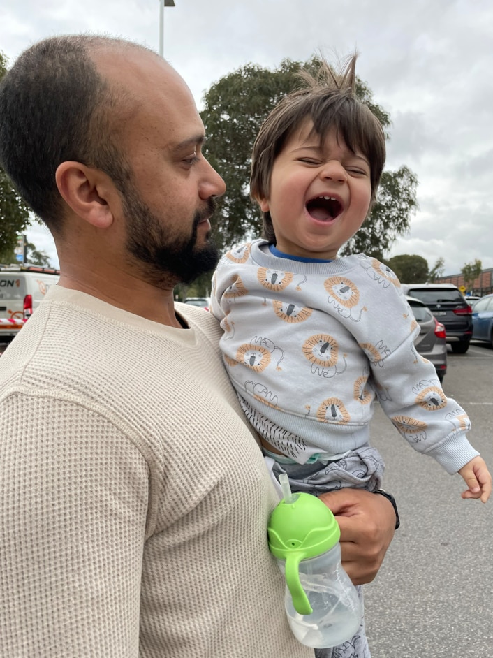 A man wearing a cream top holds a little boy smiling and wearing a blue tracksuit.