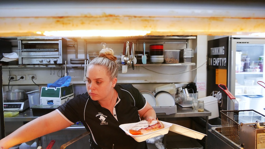 A young woman reaches across to a hot stove while packaging a takeaway meal in a styrofoam container