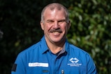 A man with moustache smiles at the camera in a blue volunteer shirt.