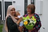 A woman in a hijab holds her baby, while an older woman holding a bunch of flowers looks on
