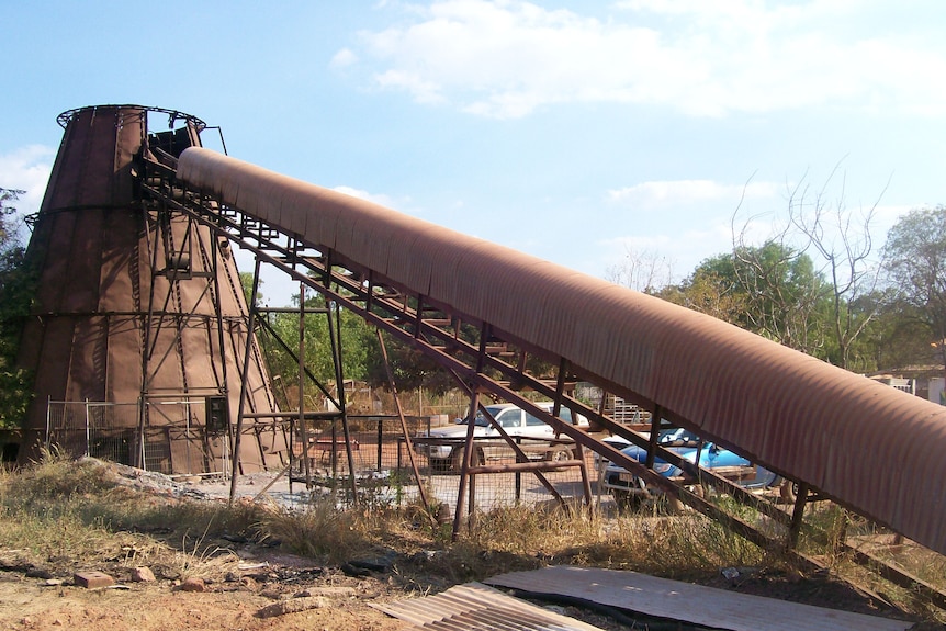 A rusted conical silo with a 30 degree conveyor from the top to the ground. Bricks and corrugated iron sheets on ground.