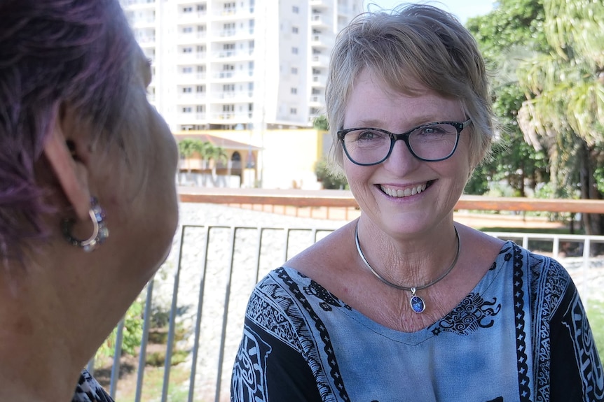 a woman with short blonde hair smiles as she looks towards another woman, sitting on a park bench