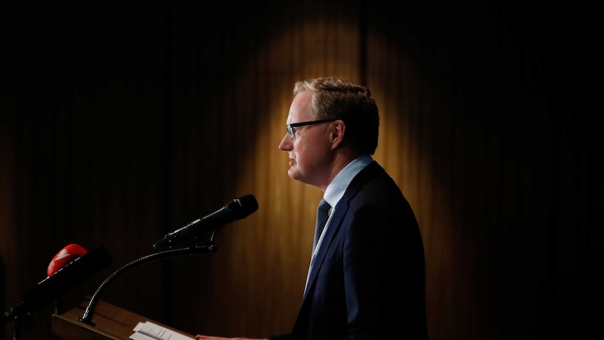 RBA Governor Phillip Lowe standing at a podium under a spotlight.