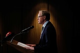 RBA Governor Phillip Lowe standing at a podium under a spotlight.