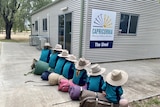A line of school children wearing large brimmed hats sitting on swags. There is a sign of the Capricornia shed facility.