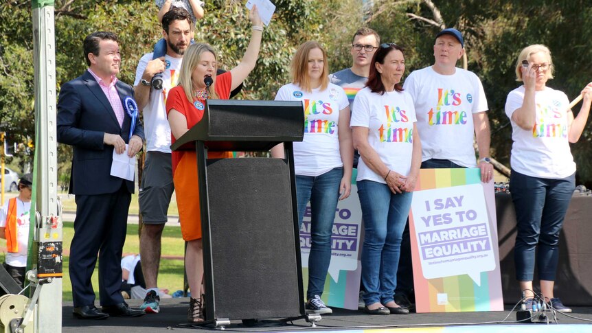 Parliamentarians stand on stage wearing rainbow "it's time" t-shirts.