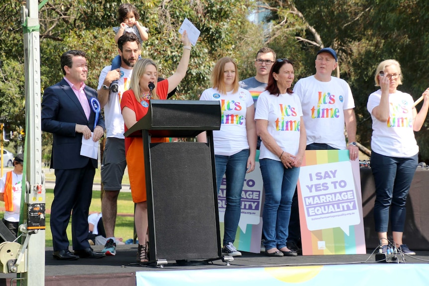 Parliamentarians stand on stage wearing rainbow "it's time" t-shirts.