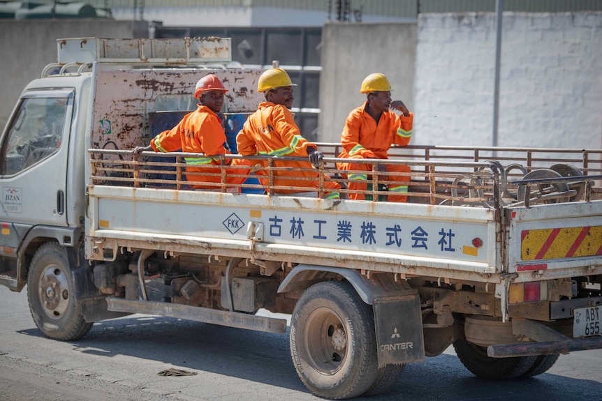 Three men wearing construction safety gear sit in the back of a work truck