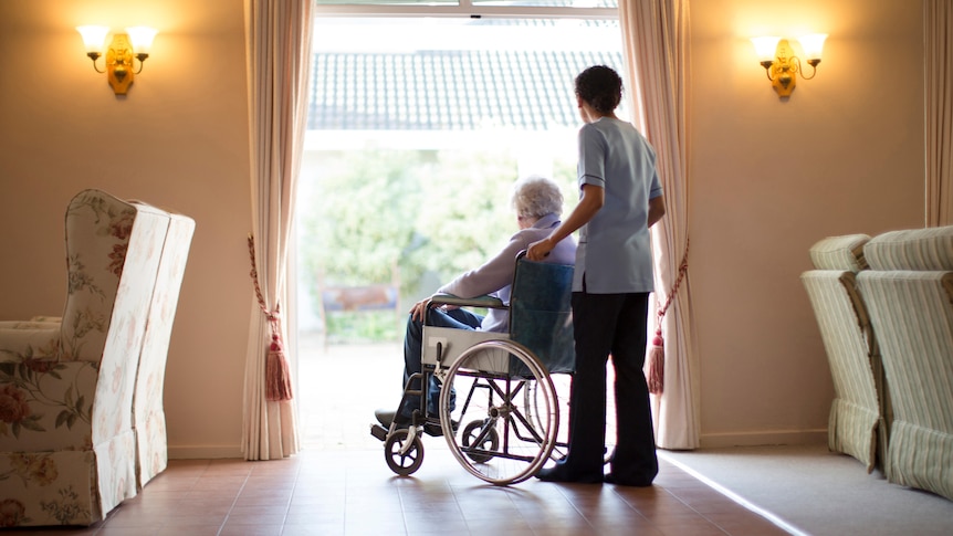 A woman in a wheelchair pushed by a man in an aged care facility.