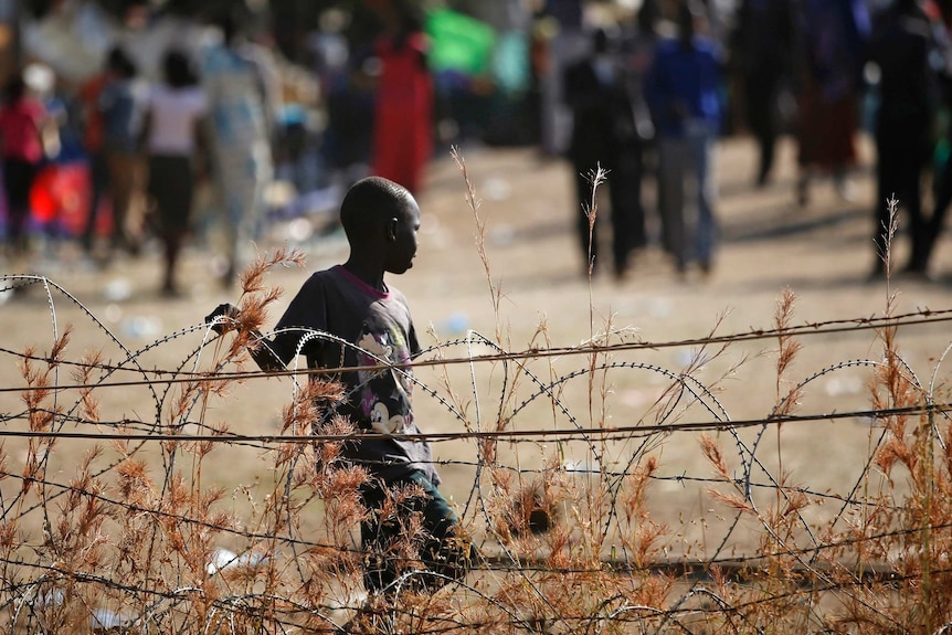 A small boy stands near a barbed wire fence with brown dry land around him.