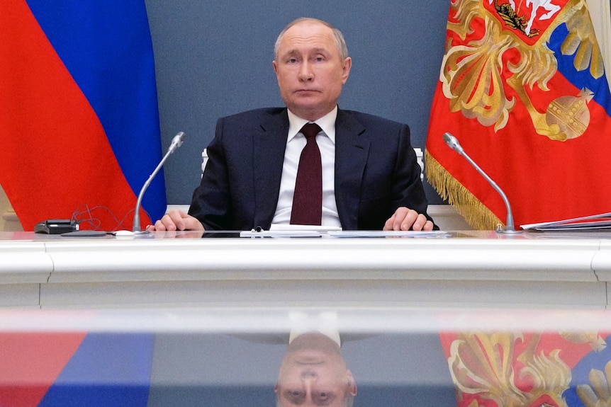 vladimir putin in a suit sits at a desk with a  microphone on it