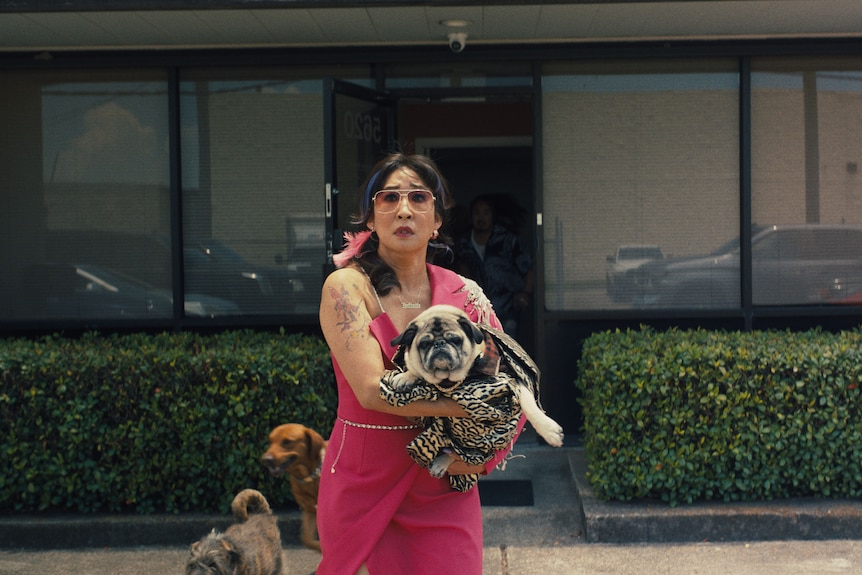 Sandra Oh in a pink jumpsuit with sleeve tattoos holds a pug and looks concerned