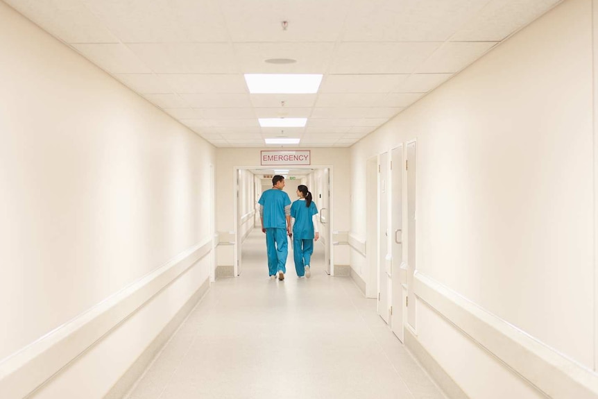 Two doctors in blue scrubs walking down a white hallway beneath an emergency sign.