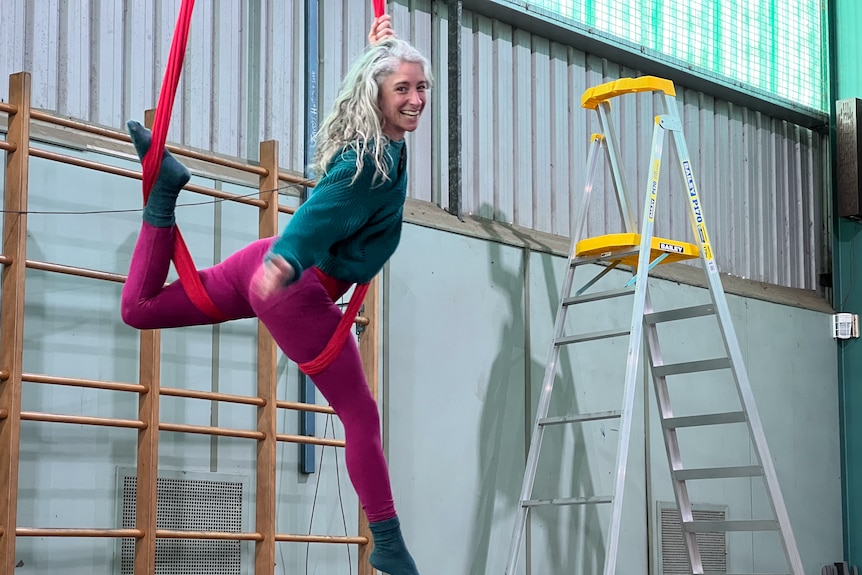 A woman with long grey hair hanging upright, with legs supported by and wound around a red aerial hammock.