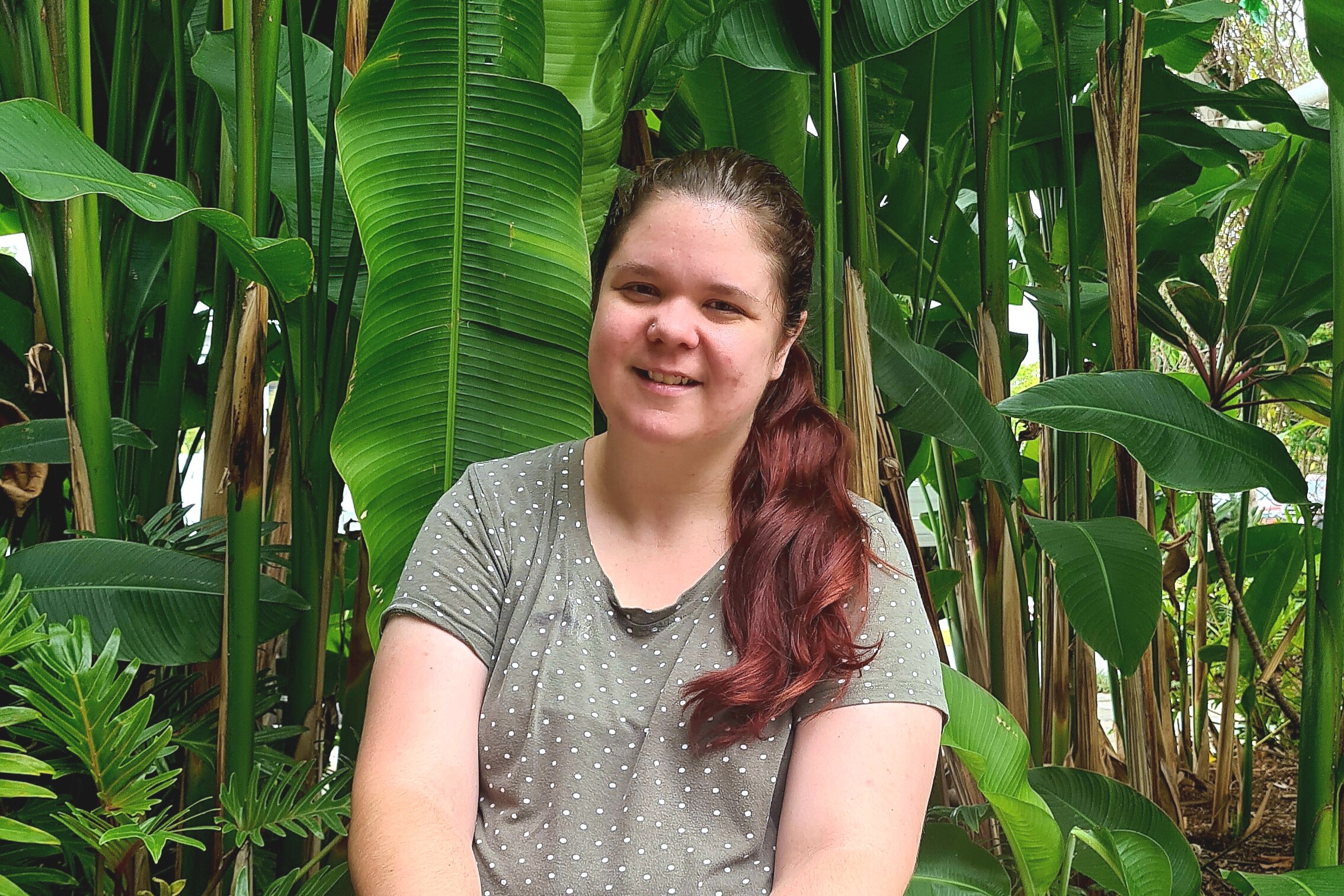 A woman wearing a grey spotted shirt smiles while standing in front of green tropical plants. 