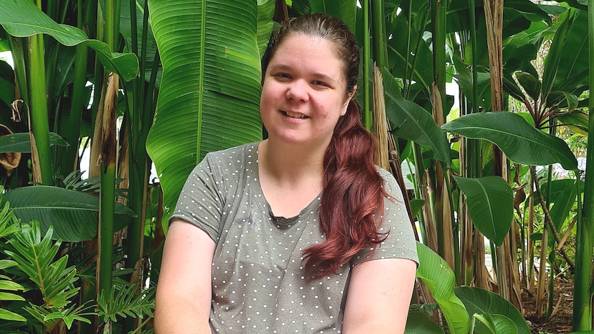 A woman wearing a grey spotted shirt smiles while standing in front of green tropical plants. 