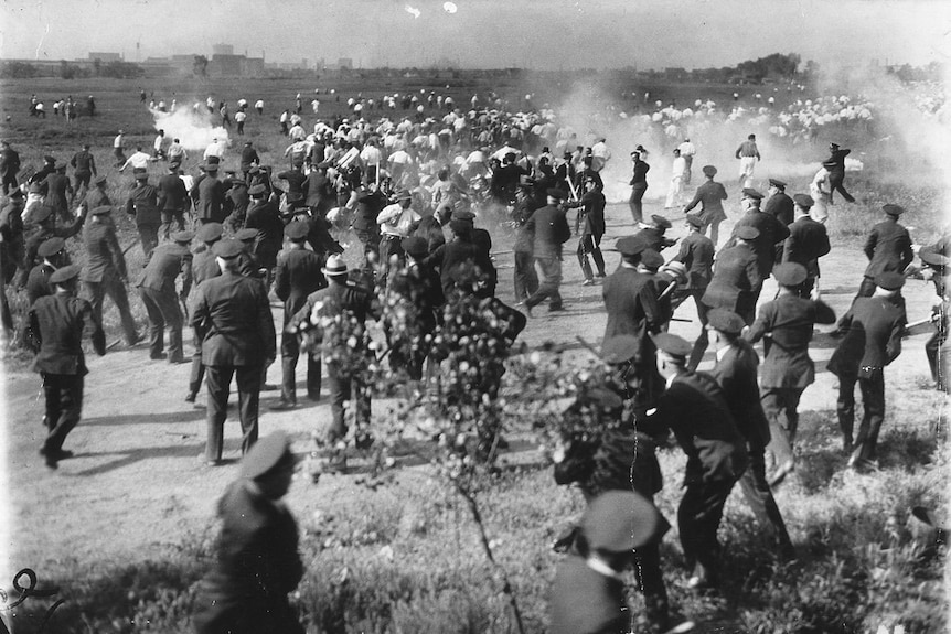 black and white image of protesters running across a field from an armed police squad, smoke from artillery fire can be seen.