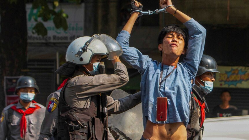 A young man in blue denim shirt raises handcuffed hands above his head as police escort him on road.