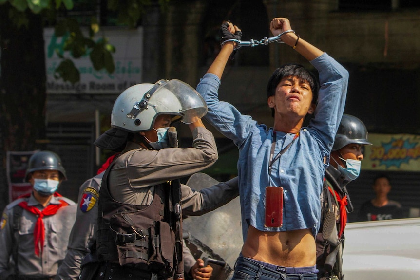 A young man in blue denim shirt raises handcuffed hands above his head as police escort him on road.