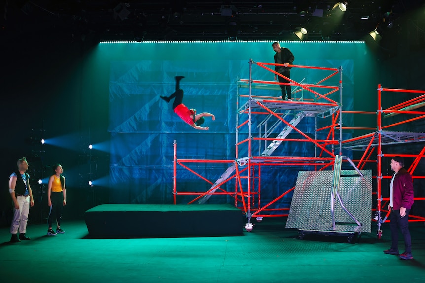 An Asian man is mid-air, having backflipped of scaffolding on a stage. Onlookers watch from the floor and scaffold.