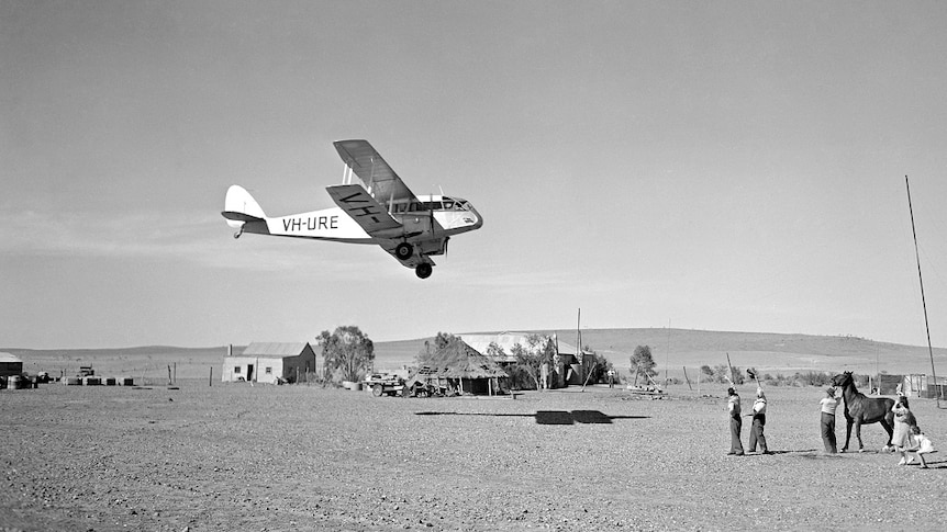 A black and white photograph of two men, a woman and a child look up at a plane flying overhead.