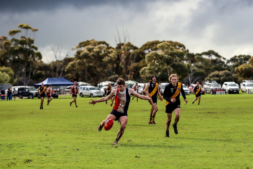 A player kicks a football as other players and spectators watch on at a country footy ground 