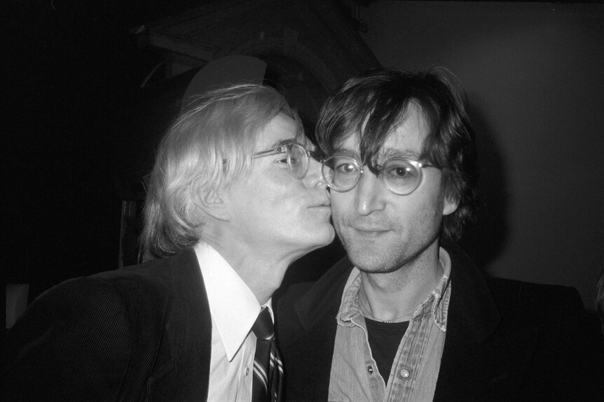 A black-and-white photograph of Andy Warhol kissing John Lennon's cheek