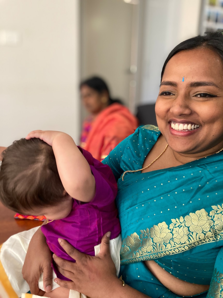 A woman in an aqua sari smiles with a baby on her lap, behind her is the blurred image of another woman