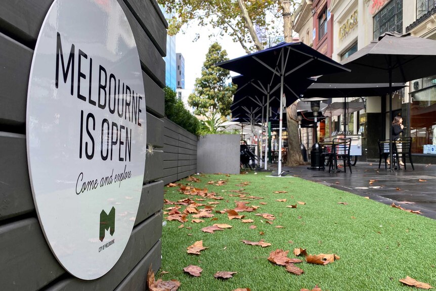A sign saying Melbourne is open, come and explore, next to artificial lawn and a cafe.