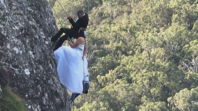 A woman in a wedding dress and a man in a tuxedo hanging from a rock above bushland.