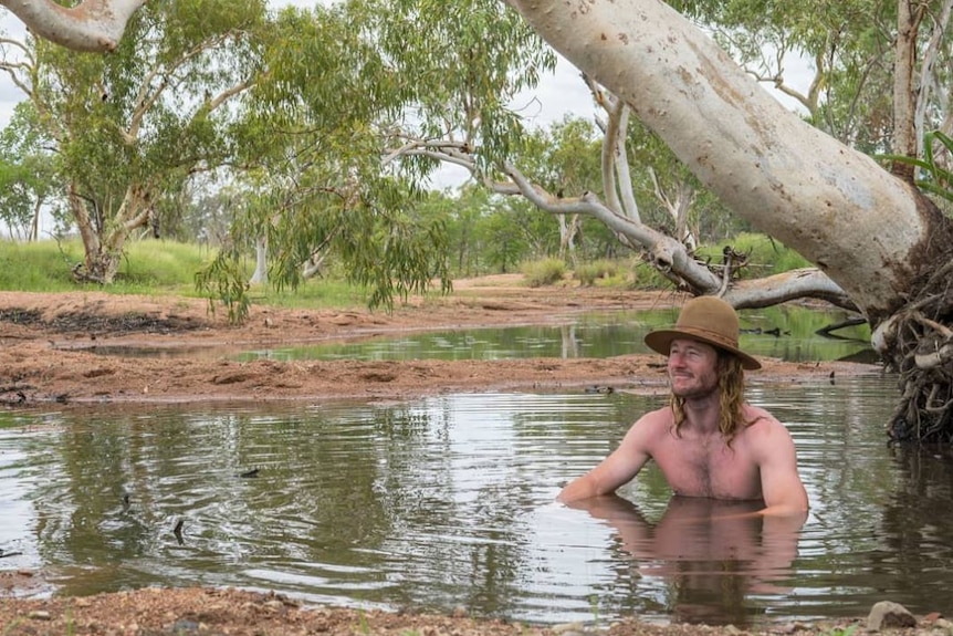 A shirtless, long-haired man wearing a hat sits in a waterhole.