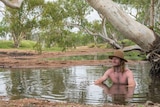 A shirtless, long-haired man wearing a hat sits in a waterhole.