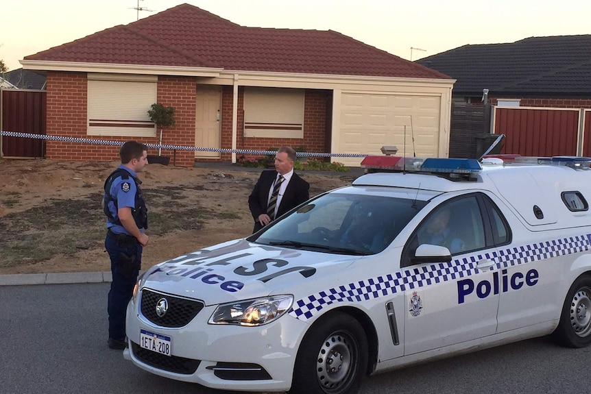 Two police officers, one a plain clothes, stand next to a police car outside a house with police tape surrounding it.