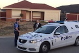 Two police officers, one a plain clothes, stand next to a police car outside a house with police tape surrounding it.