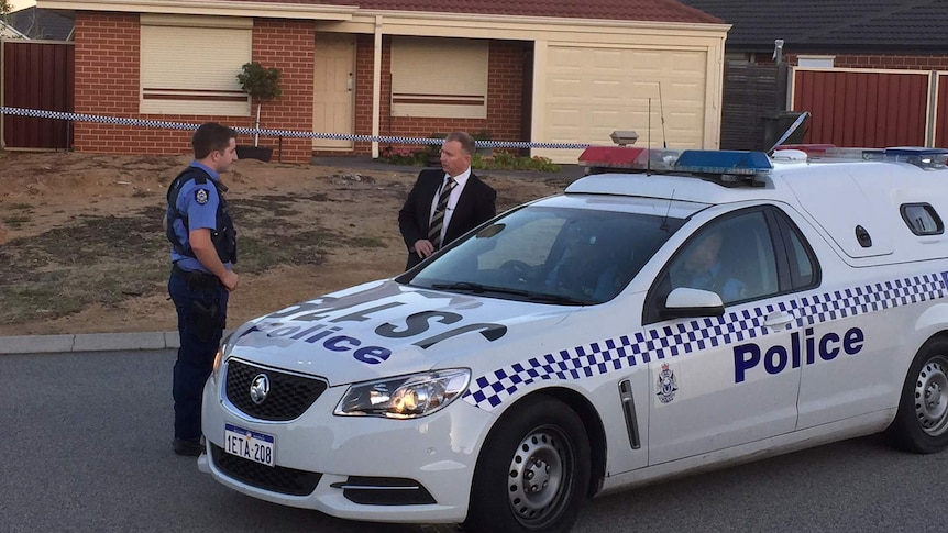 Two police officers, one a plain clothes, stand next to a police car outside a house with police tape surrounding it.