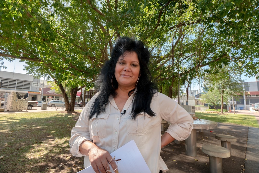 A woman holds her glasses and paper and stands under a large shady tree.