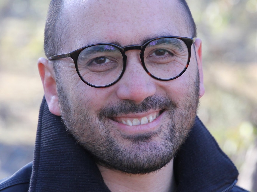 A man with round glasses, short hair and stubble smiles at the camera.