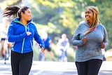 Two women in active wear smile at each other as they run in a park.